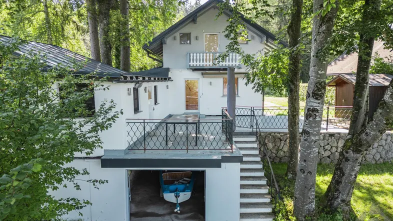 Weißes Haus mit Balkon, Terrasse und integrierter Bootsgarage, umgeben von hohen Bäumen.