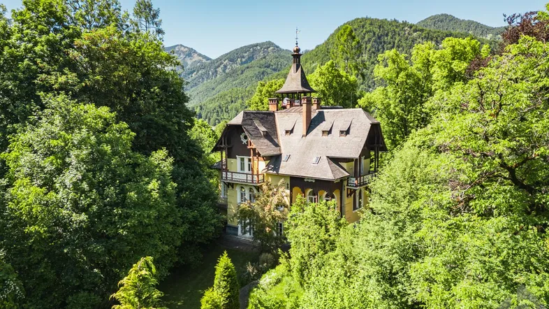Eine prächtige gelbe Villa mit markantem Turm, umgeben von üppigem Grün und majestätischen Bergen.