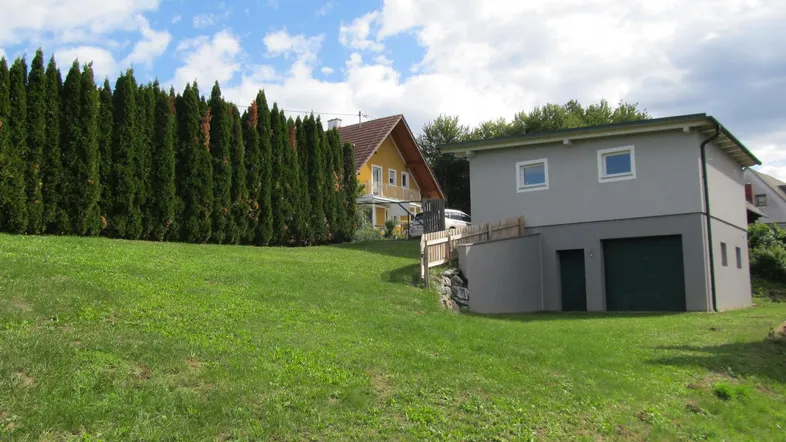 Graues Gebäude mit grüner Garage auf einem grasbewachsenen Hang, umgeben von Bäumen und einem hohen Zaun.