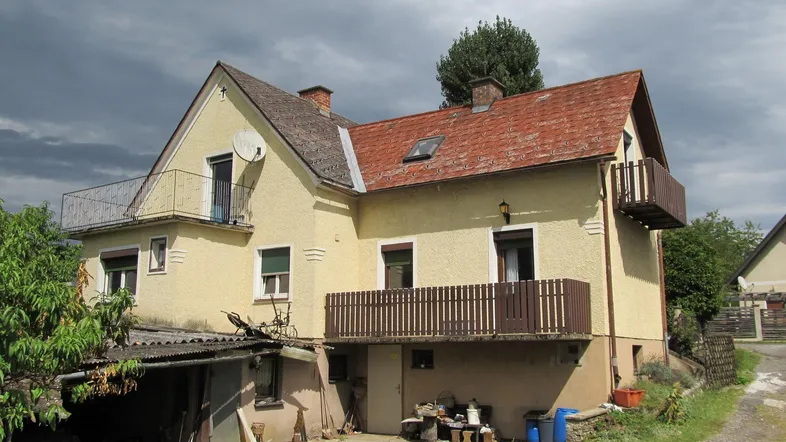 Gelbes Einfamilienhaus mit rotem Ziegeldach, zwei Balkonen und einfacher Außenanlage unter bewölktem Himmel.