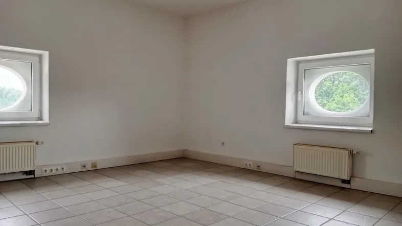 Heller, leerer Raum mit Fliesenboden und zwei markanten runden Fenstern, ideal als Bürofläche.