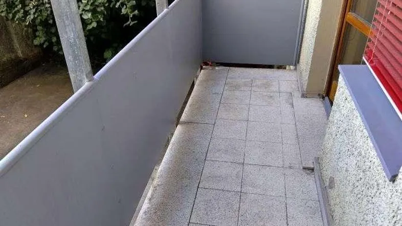 Langer, schmaler Balkon mit Fliesenboden und Blick auf grüne Bäume im Hintergrund.