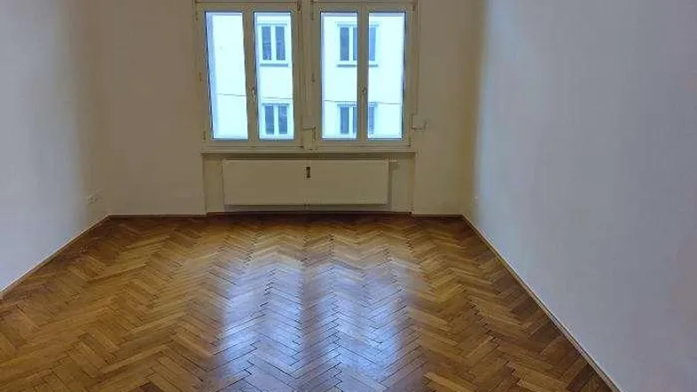Helles Zimmer mit klassischem Fischgrätparkettboden und großen Fenstern, die viel Tageslicht hereinlassen.