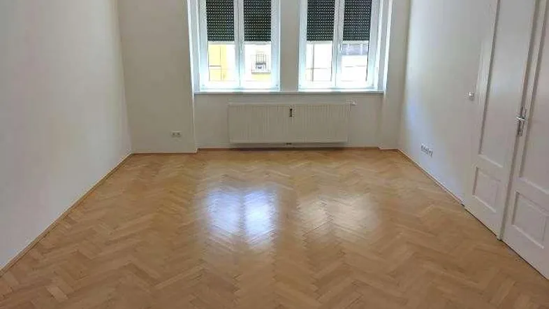 Geräumiges Zimmer mit Fischgrätparkett, weißen Wänden und zwei Fenstern mit Jalousien.