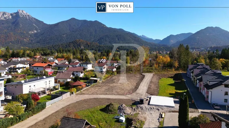 Luftbild eines Baugebiets mit vorbereiteten Grundstücken und beeindruckendem Bergpanorama im Hintergrund.