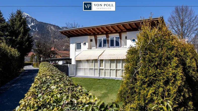 Modernes Einfamilienhaus mit gepflegtem Vorgarten, Hecke und Bergblick unter blauem Himmel.