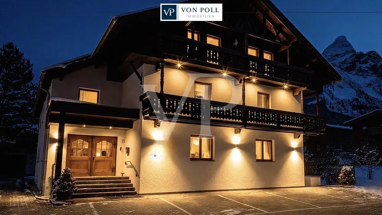 Beleuchtetes Chalet-Haus in den Bergen bei Nacht mit traditionellen Holzbalkonen und verschneiter Umgebung.