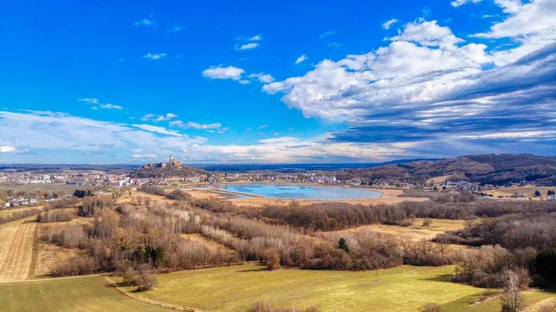 Weitläufige Landschaft mit See, Feldern und einer Burg in der Ferne unter blauem Himmel.