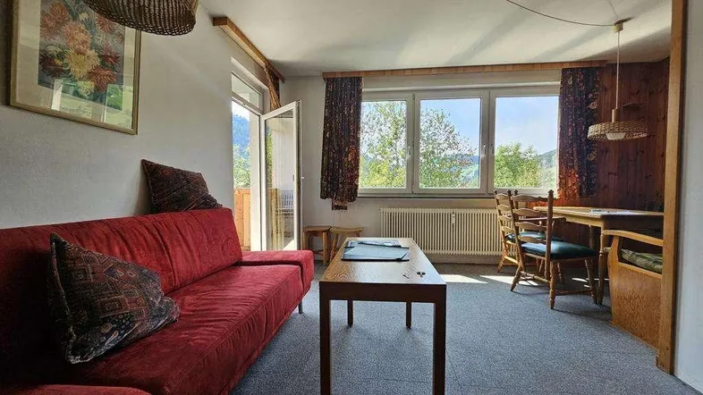 Gemütliches Wohnzimmer mit rotem Sofa, Esstisch und Zugang zum Balkon mit Bergblick.