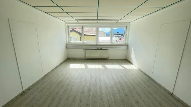 Heller Büroraum mit Laminatboden, abgehängter Decke und Fenstern mit Bergblick.