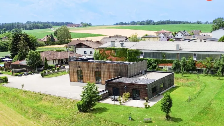 Luftaufnahme eines modernen Gebäudekomplexes mit Holzfassade, umgeben von grünen Feldern und ländlicher Bebauung.