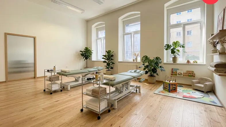 Heller Therapieraum mit zwei Behandlungsliegen, großen Fenstern und einem kleinen Bereich für Kinder. Der Raum hat einen schönen Holzboden.