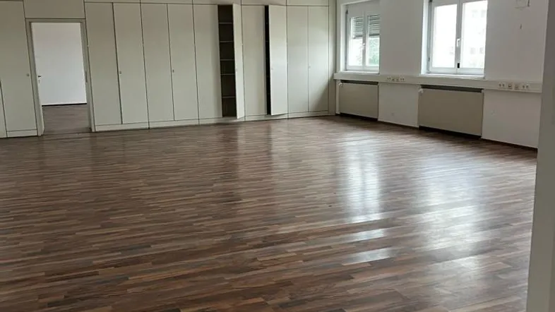 Großer, leerer Büroraum mit Laminatboden, Einbauschränken und Fenstern, die viel Tageslicht hereinlassen.