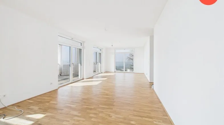 Helles, leeres Wohnzimmer mit Parkettboden und Zugang zu einem Balkon.