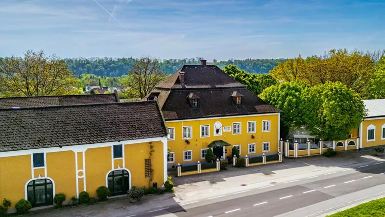 Luftaufnahme eines gelben Gebäudekomplexes mit dunklen Dächern und einer Straße davor unter blauem Himmel.