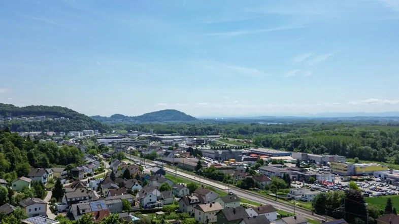 Weitläufige Luftaufnahme einer Siedlung mit viel Grün und Hügellandschaft im Hintergrund unter blauem Himmel.