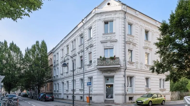 Weißes Eckgebäude mit klassischer Fassade und kleinem Balkon an einer städtischen Straße mit Bäumen.