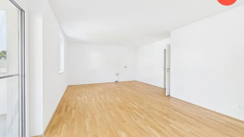 Helles, leeres Zimmer mit Parkettboden und großem Fenster, das viel Tageslicht hereinlässt.