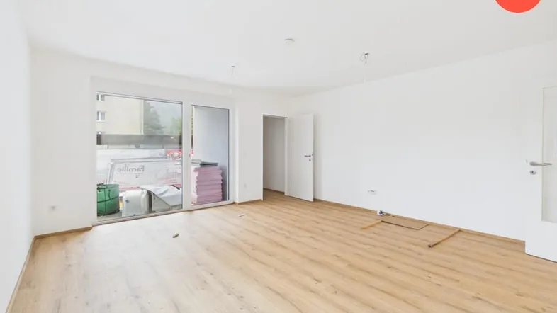 Helles, leeres Wohnzimmer mit Laminatboden und Zugang zu einem Balkon in einem Neubau.