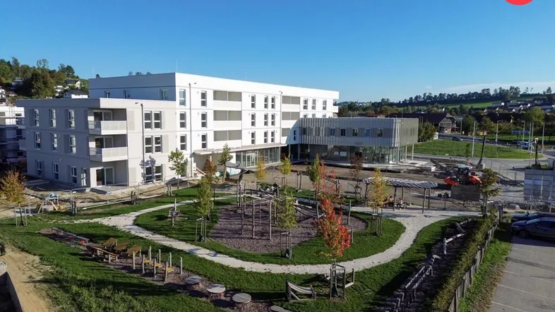 Luftaufnahme eines modernen Neubaus mit angrenzendem Spielplatz und Grünflächen unter blauem Himmel.
