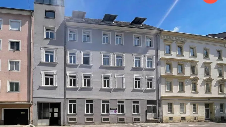 Gebäudefassade eines Mehrfamilienhauses mit grauer Putzfassade, weißen Fenstern und Solarpaneelen auf dem Dach.