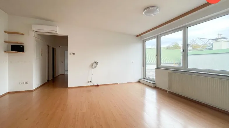 Helles, leeres Wohnzimmer mit Holzfußboden und großen Fenstern, die viel Tageslicht hereinlassen.