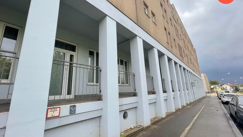 Langes Wohngebäude mit mehreren Loggien und Balkonen, davor ein Gehweg und parkende Autos unter bewölktem Himmel.