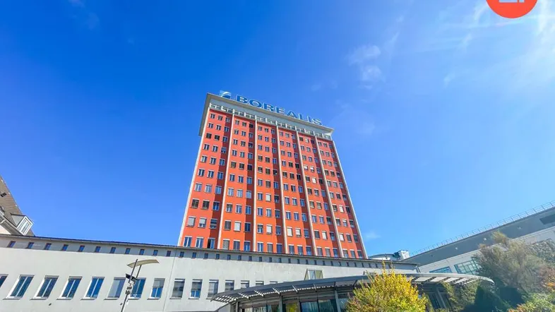 Hohes Gebäude mit markanter orangeroter Fassade und blauer Himmel, umgeben von niedrigeren Gebäuden und Grünflächen.