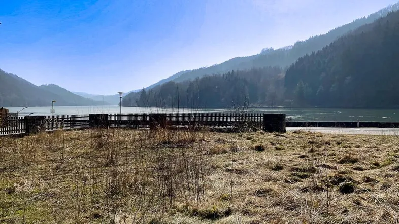 Weitläufiger Blick auf einen See oder Fluss, umgeben von bewaldeten Bergen unter blauem Himmel.