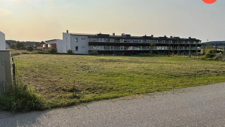 Ausgedehntes, unbebautes Grundstück mit Gras und einer Straße, dahinter ein mehrstöckiges Wohngebäude.