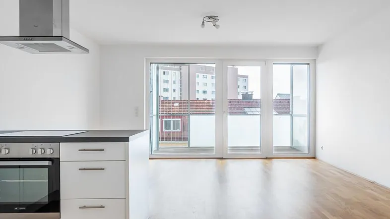 Heller Wohnbereich mit offener Küche, Holzboden und großen Fenstern zum Balkon.