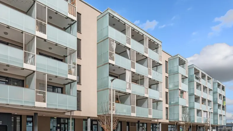 Ansicht der modernen Wohnanlage mit zahlreichen Balkonen, Geschäften im Erdgeschoss und einer Rampe.
