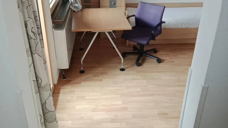 Helles Schlafzimmer mit Laminatboden, Schreibtisch und Kleiderschrank, durch einen Torbogen sichtbar.