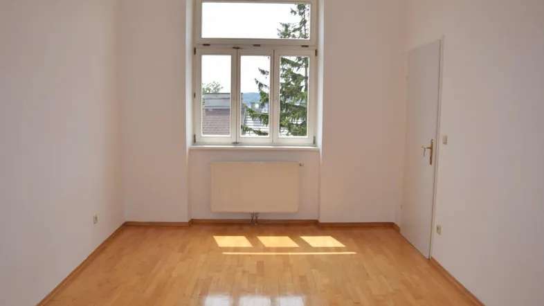Helles Zimmer mit Parkettboden und großem Fenster, das viel Tageslicht hereinlässt.
