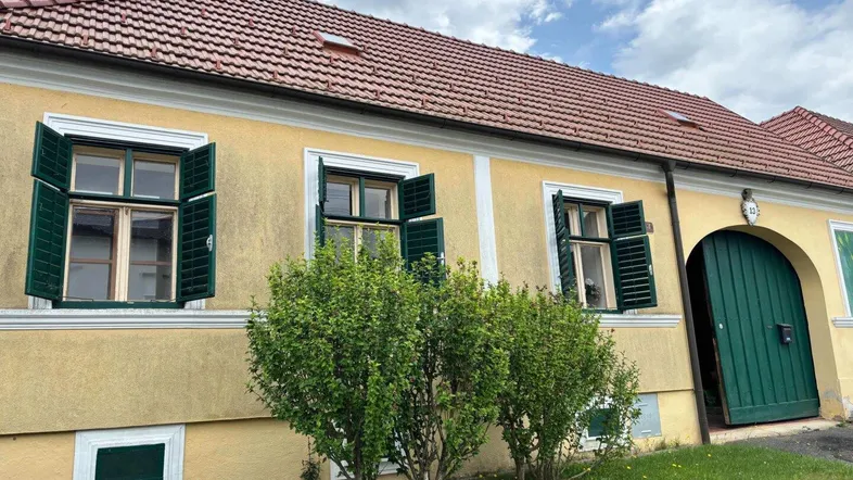 Traditionelles gelbes Haus mit roten Ziegeln und grünen Fensterläden unter bewölktem Himmel.