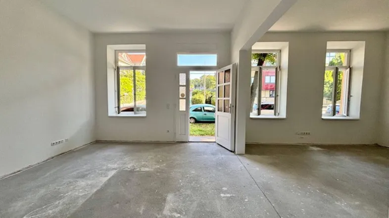 Leerer Raum in Renovierung mit Betonboden und weißen Wänden, viel Tageslicht durch Fenster.
