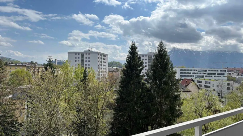 Weitblick vom Balkon über die Stadt mit grünen Bäumen und Bergen im Hintergrund.