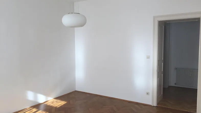 Helles Zimmer mit Fischgrätparkett und weißen Wänden, durch eine offene Tür sichtbar.