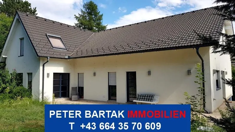 Weißes Einfamilienhaus mit dunklem Dach, kleiner Terrasse und Gartenbereich unter blauem Himmel.
