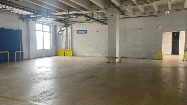 Großer, leerer Industrieraum mit Betonboden, hohen Decken und sichtbaren Installationen, ideal für Lagerung.