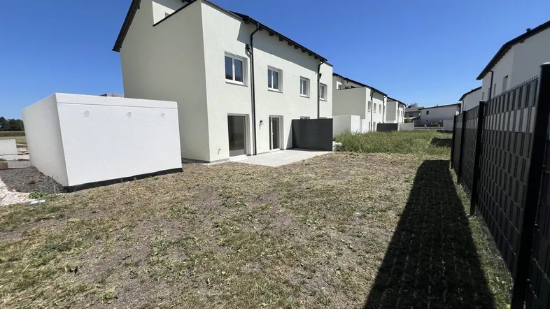Neubau-Reihenhaus mit weißer Fassade, kleiner Terrasse und noch ungestaltetem Gartenbereich unter blauem Himmel.