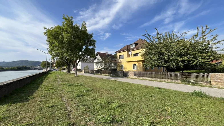 Blick auf die Straße entlang des Flusses mit Wohnhäusern und viel Grün unter blauem Himmel.