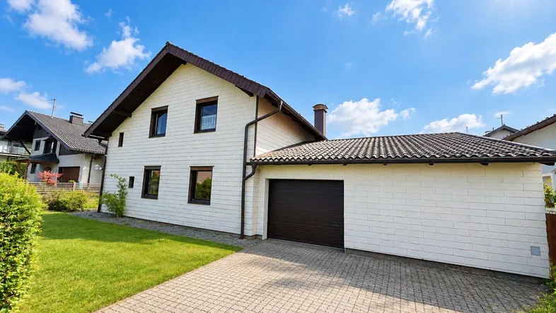 Weißes Einfamilienhaus mit Satteldach, integrierter Garage und gepflasterter Auffahrt unter blauem Himmel.