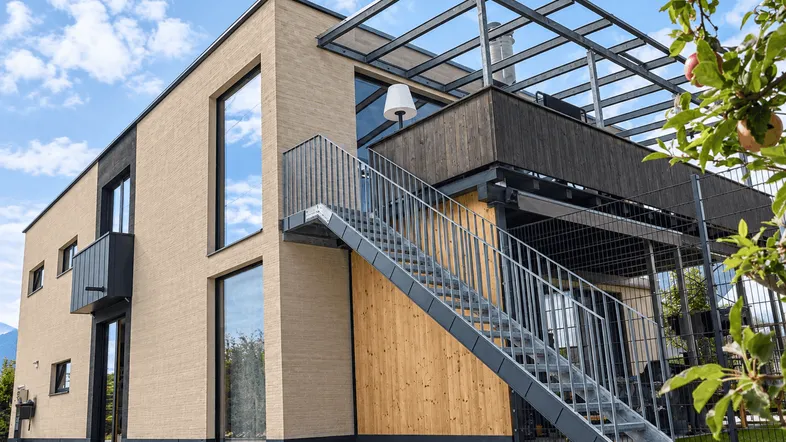 Modernes Wohnhaus mit beiger Klinkerfassade, großen Fenstern und Außentreppe zur Dachterrasse mit Pergola.