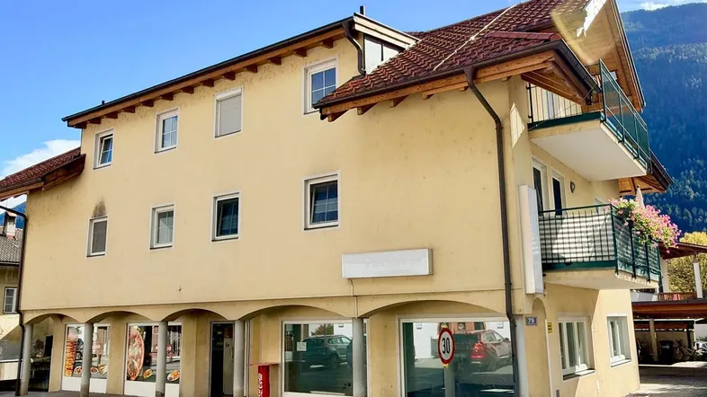 Gebäudeansicht mit Geschäften im Erdgeschoss und Wohnungen darüber, umgeben von Bergen.