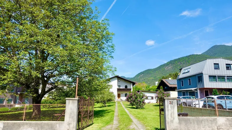 Einfahrt zu einem Wohnhaus mit großem Garten, umgeben von Bäumen und Bergen im Hintergrund.