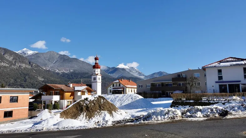 Panoramablick auf eine verschneite Wohnsiedlung mit Kirche und majestätischen Bergen im Hintergrund unter blauem Himmel.