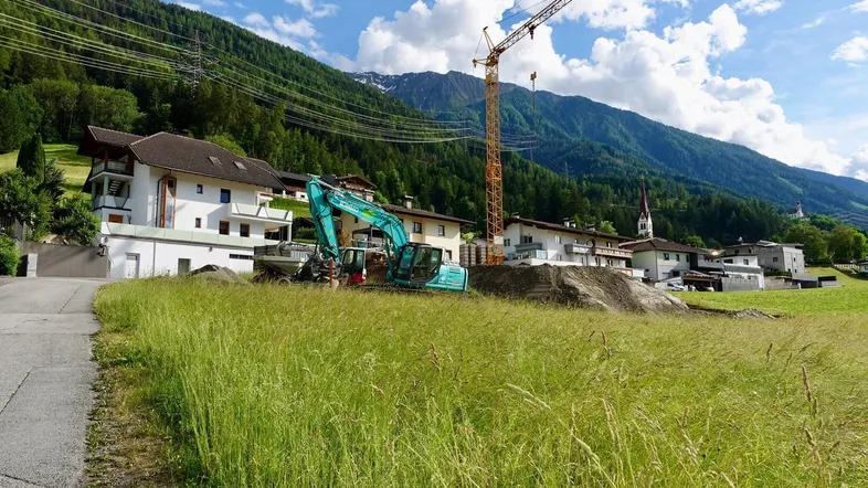 Baustelle mit Bagger und Kran vor einer Bergkulisse und Wohnhäusern in ländlicher Umgebung.