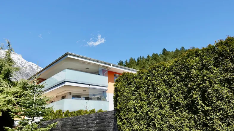 Modernes Gebäude mit Balkonen und Glasbrüstungen, umgeben von grünen Bäumen und Hecken.