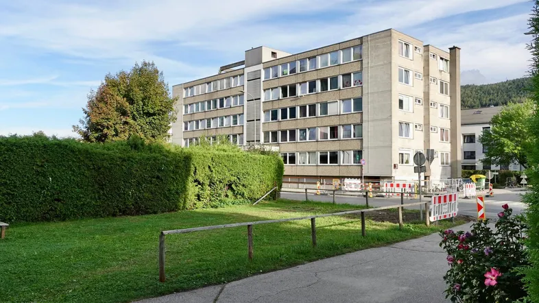Mehrstöckiges Wohngebäude mit Betonfassade, umgeben von Grünflächen und Hecken, mit Bergblick im Hintergrund.